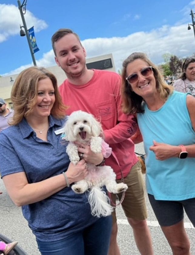 Three people smile for a photo outdoors on a sunny day, with a small white dog in the arms of the woman on the left. Other people and blue sky with clouds are visible in the background.