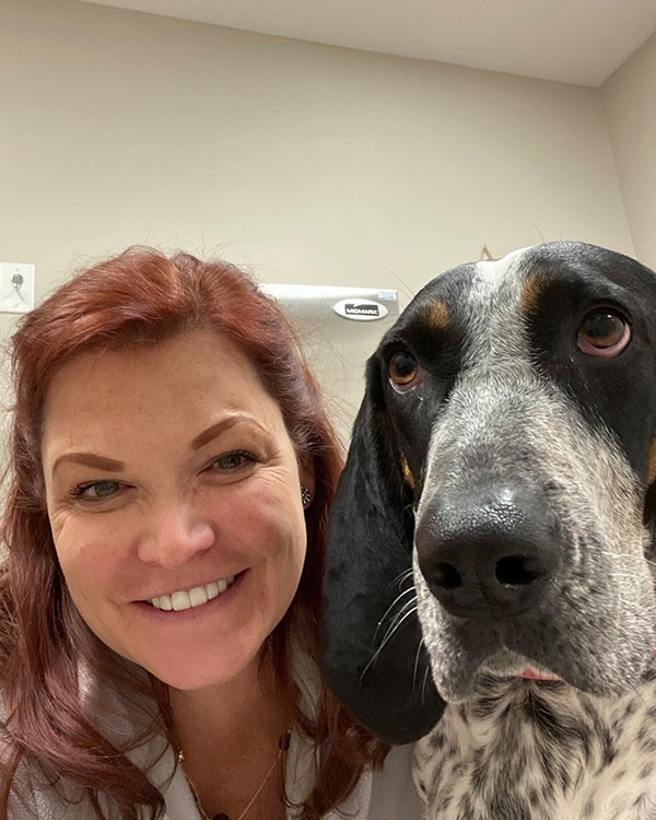 A smiling woman with red hair poses for a selfie next to a black and white dog with long ears in an indoor setting.