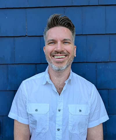 Smiling man with short brown hair and a beard, wearing a white short-sleeve button-up shirt, standing in front of a blue wall with horizontal panels.