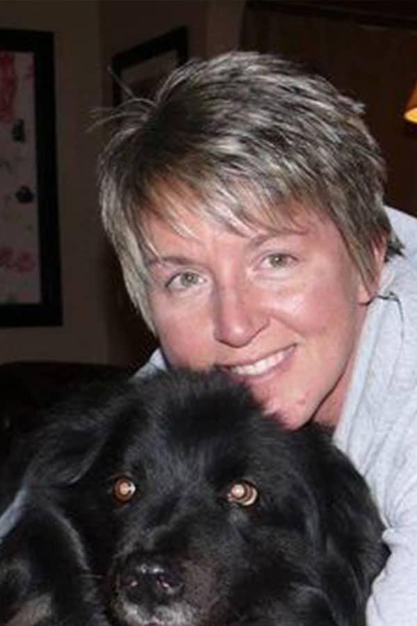 A woman with short, light brown hair smiles while hugging a large, fluffy black dog indoors.