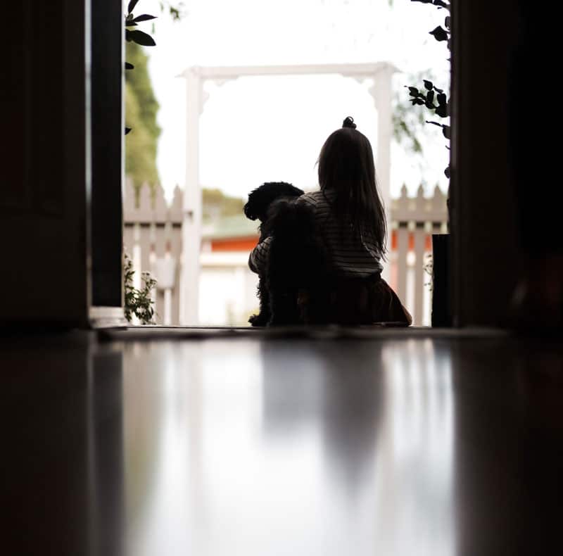 A small child sits at an open doorway, facing outside, and hugs a black dog. The scene is softly lit, with reflections on the floor and a white picket fence visible in the background.